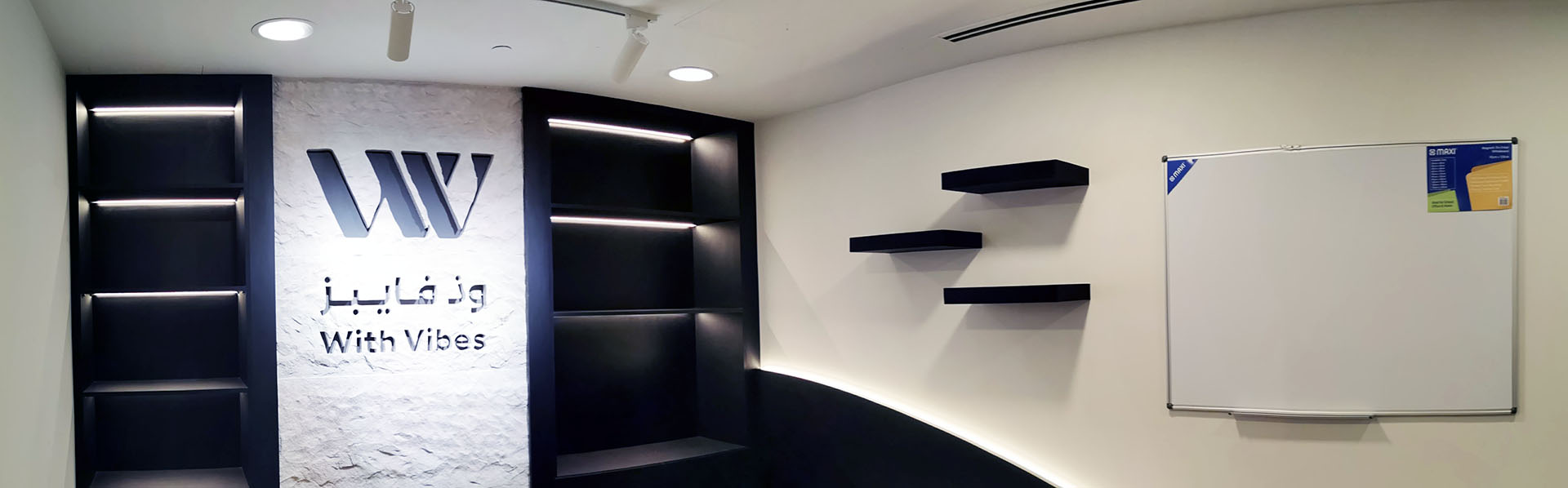 With Vibes office fit-out in Lusail with Tufa stone logo wall, black cabinetry, floating shelves, whiteboard, and integrated lighting